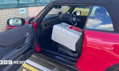 The side exterior of a red convertible Mini Cooper car parked in a car park. The passenger side door is open with a large grey box on the seat.