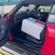 The side exterior of a red convertible Mini Cooper car parked in a car park. The passenger side door is open with a large grey box on the seat.