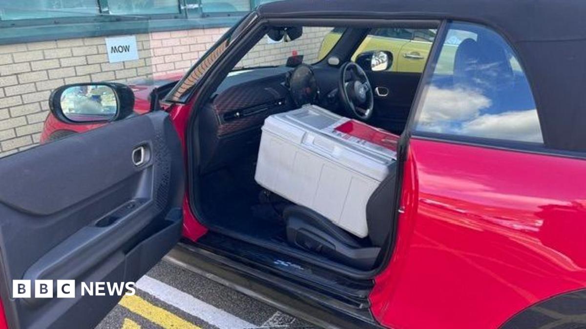 The side exterior of a red convertible Mini Cooper car parked in a car park. The passenger side door is open with a large grey box on the seat.