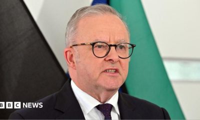 Australia's Prime Minister Anthony Albanese speaks at a news conference in front of several flags. He is wearing a dark suit with a red spotted tie and dark glasses