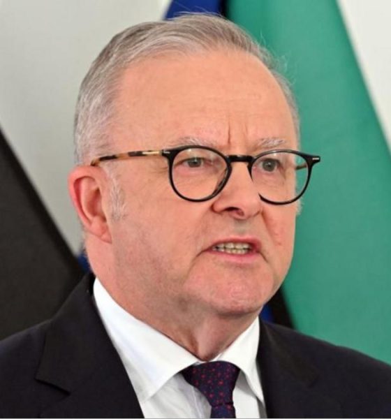 Australia's Prime Minister Anthony Albanese speaks at a news conference in front of several flags. He is wearing a dark suit with a red spotted tie and dark glasses