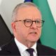 Australia's Prime Minister Anthony Albanese speaks at a news conference in front of several flags. He is wearing a dark suit with a red spotted tie and dark glasses