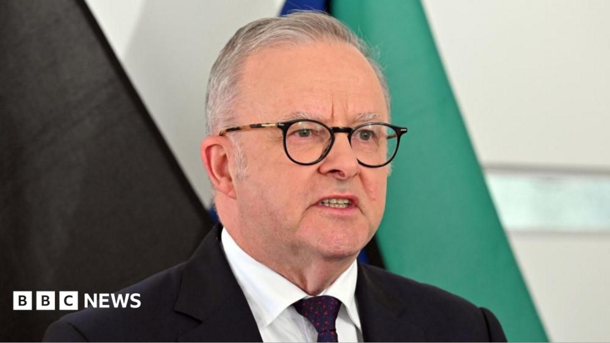 Australia's Prime Minister Anthony Albanese speaks at a news conference in front of several flags. He is wearing a dark suit with a red spotted tie and dark glasses