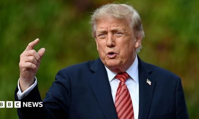 Donald Trump gestures to someone off camera. He is wearing a navy blue suit jacket, white shirt and a reddish-striped tie.