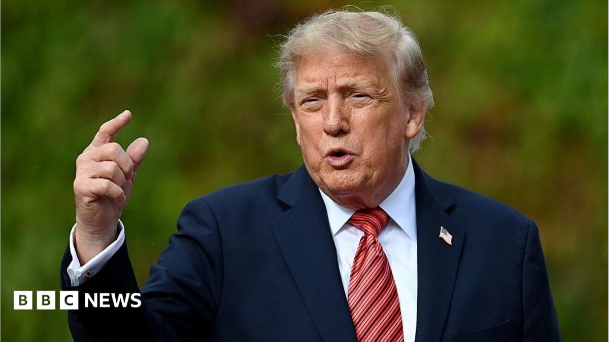 Donald Trump gestures to someone off camera. He is wearing a navy blue suit jacket, white shirt and a reddish-striped tie.
