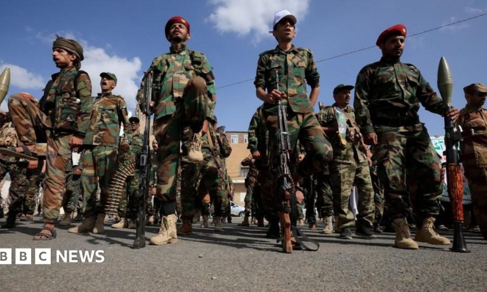 A dozen Houthi men wearing combat clothes, berets and holding large guns. The sky is blue and the men are looking straight ahead.