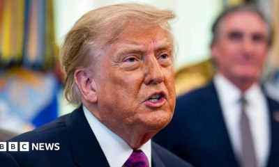 Donald Trump speaks, he is wearing a blue suit jacket, white shrit with a red and purple tie. The background is blurred.