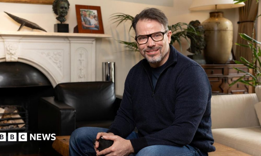 Chris Rokos sitting in a room wearing a dark blue jumper and blue jeans. In the background there is a fireplace, sofa and chest of drawers.