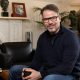 Chris Rokos sitting in a room wearing a dark blue jumper and blue jeans. In the background there is a fireplace, sofa and chest of drawers.