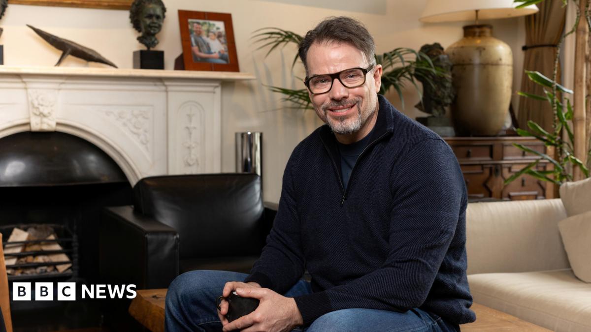 Chris Rokos sitting in a room wearing a dark blue jumper and blue jeans. In the background there is a fireplace, sofa and chest of drawers.