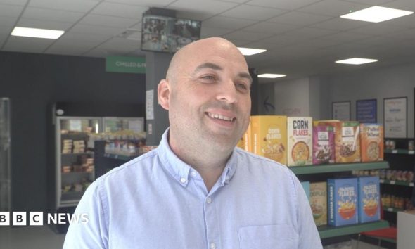 Stephen Brindley smiling and looking to the side of the frame. Supermarket shelves and fridges with goods including tins, cereal, and frozen meals are visible.