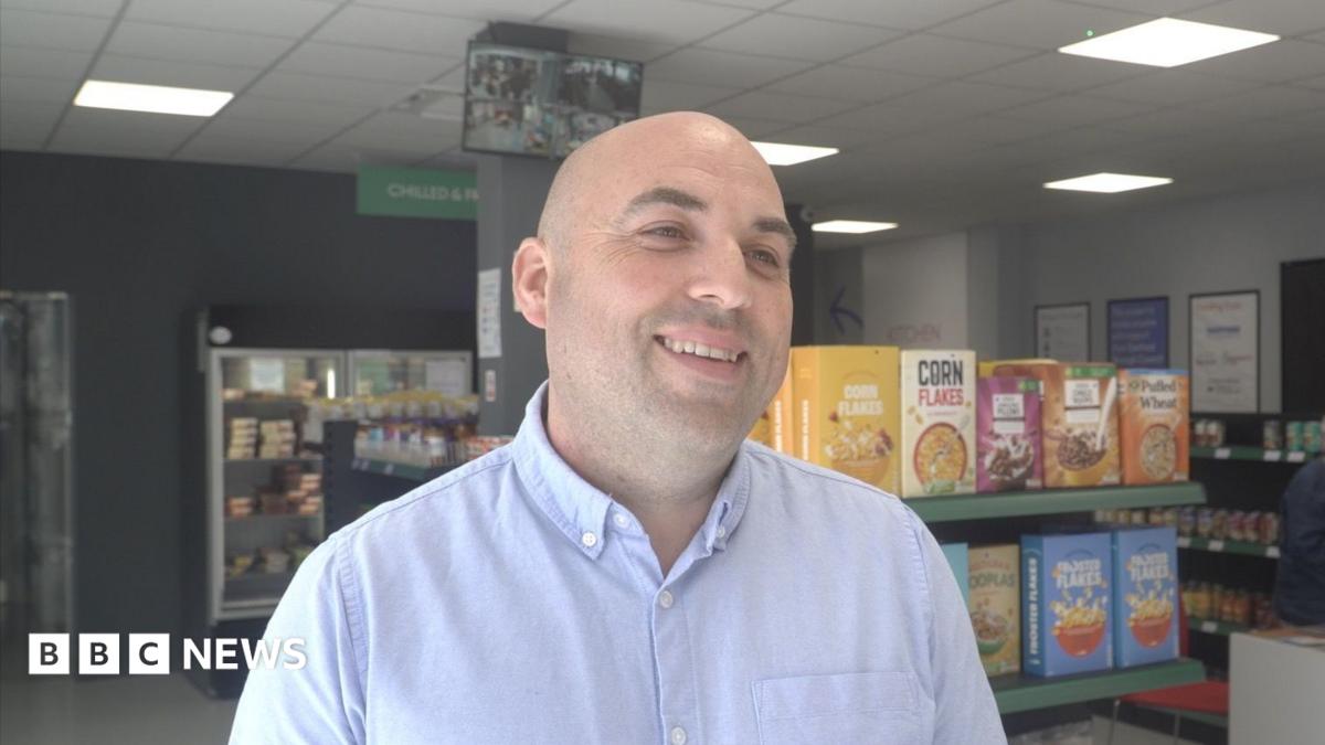 Stephen Brindley smiling and looking to the side of the frame. Supermarket shelves and fridges with goods including tins, cereal, and frozen meals are visible.