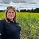 Farmer Debbie Wilkins is pictured standing in a field full of yellow flowers on a cloudy but sunny day. She has long blonde hair and a pair of sunglasses is perched on her head. She is wearing a navy blue polo shirt.