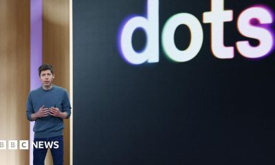 OpenAI CEO Sam Altman standing on stage at his company's annual developer conference in front of a screen with "dots" displayed behind it.