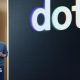 OpenAI CEO Sam Altman standing on stage at his company's annual developer conference in front of a screen with "dots" displayed behind it.