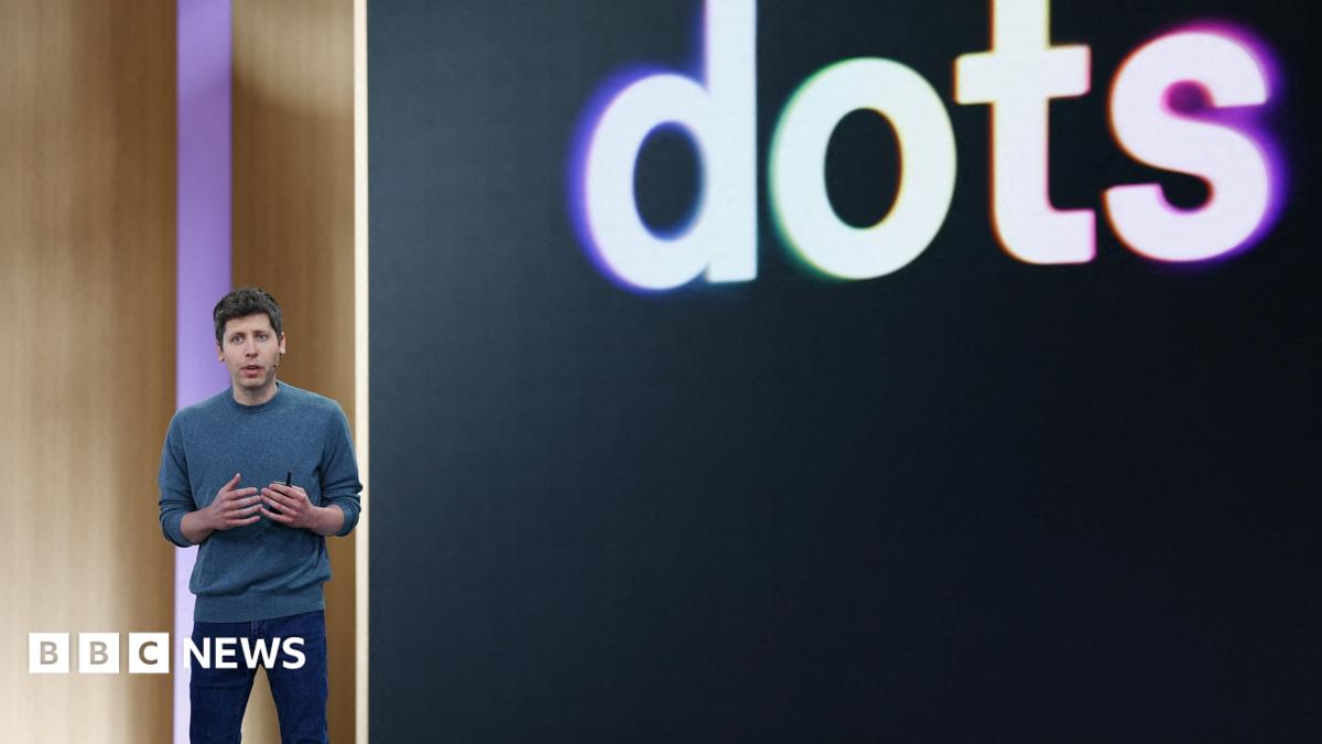 OpenAI CEO Sam Altman standing on stage at his company's annual developer conference in front of a screen with "dots" displayed behind it.