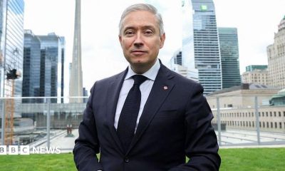 Champagne wears a suit and tie while he stands on a lawn in front of the Toronto city skyline of high-rise buildings.