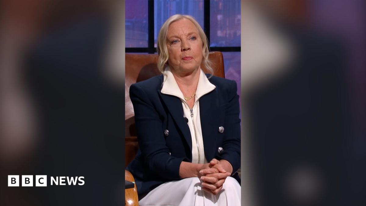 Deborah Meaden has blonde hair, She is wearing a white shirt and trousers with a dark blue jacket.