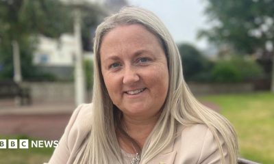 A woman called Lucy Stephenson. She is sat on a bench in a park. She has long, straight blonde hair and is wearing a cream suit.