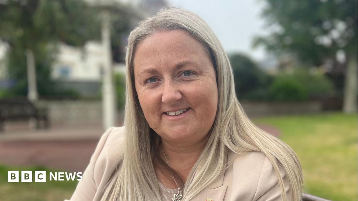 A woman called Lucy Stephenson. She is sat on a bench in a park. She has long, straight blonde hair and is wearing a cream suit.