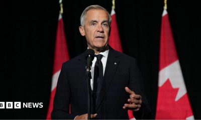 Mark Carney, in a black suit and tie gestures as he stands in front of a microphone. Behind him, against a dark background, are three red and white Canadian flags in a row.