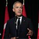 Mark Carney, in a black suit and tie gestures as he stands in front of a microphone. Behind him, against a dark background, are three red and white Canadian flags in a row.