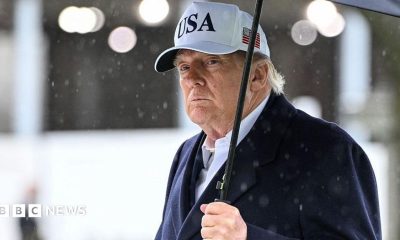 President Trump holds an umbrella as rain drops fall around him. He wears a dark suit and a white baseball cap with USA written on the front and an American flag on the side.