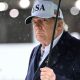 President Trump holds an umbrella as rain drops fall around him. He wears a dark suit and a white baseball cap with USA written on the front and an American flag on the side.