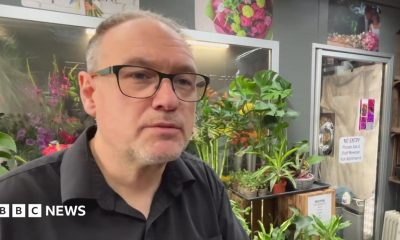 The image shows a person wearing glasses and a dark shirt standing inside what appears to be a florist or plant shop, surrounded by a large number of indoor plants and flowers. Behind the person are shelves displaying pots, bowls and glass vases.