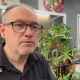 The image shows a person wearing glasses and a dark shirt standing inside what appears to be a florist or plant shop, surrounded by a large number of indoor plants and flowers. Behind the person are shelves displaying pots, bowls and glass vases.
