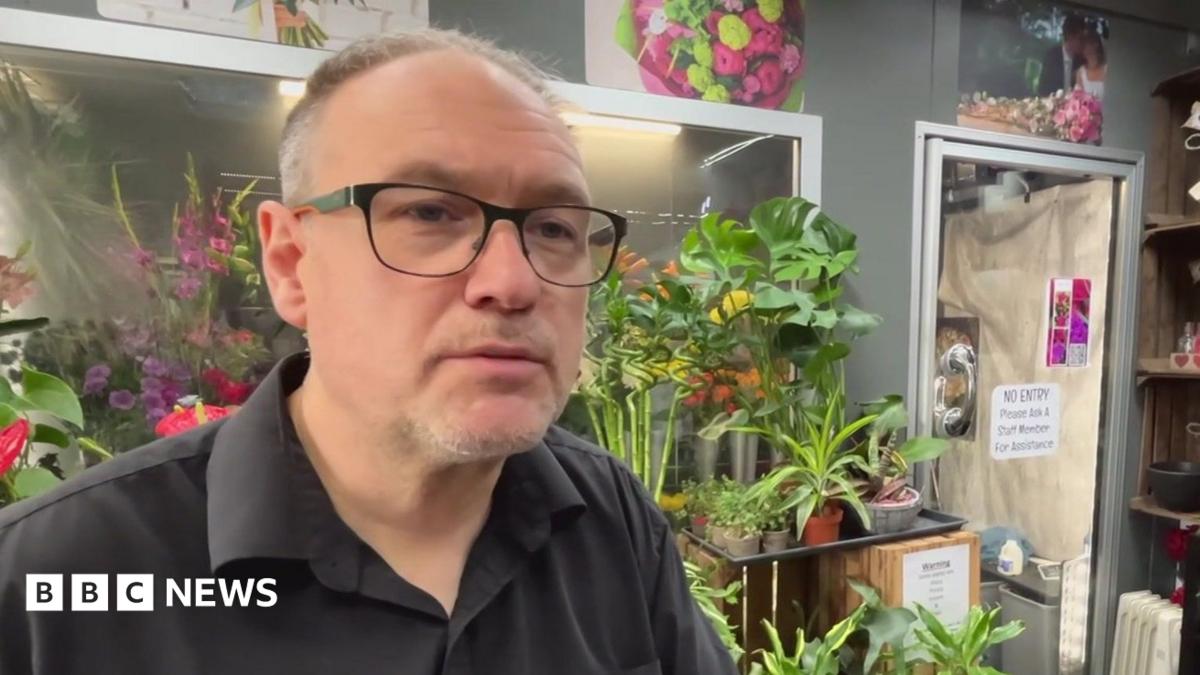 The image shows a person wearing glasses and a dark shirt standing inside what appears to be a florist or plant shop, surrounded by a large number of indoor plants and flowers. Behind the person are shelves displaying pots, bowls and glass vases.