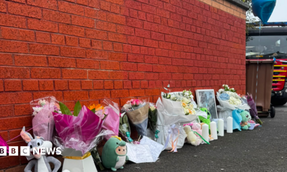 Image shows a number of floral tributes and cuddly toys left along a wall, with a fire engine in the background.