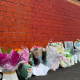 Image shows a number of floral tributes and cuddly toys left along a wall, with a fire engine in the background.