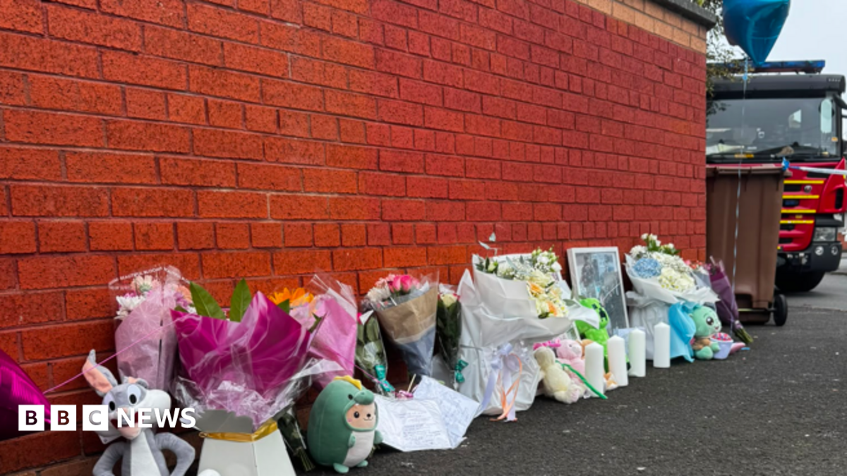 Image shows a number of floral tributes and cuddly toys left along a wall, with a fire engine in the background.