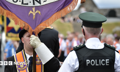 A man is wearing an orange sash and a black bowler hat and holding a banner. A policeman has his back to the camera. He has a black vest on stating police in white lettering. He has a cap and a white shirt on. A parade is out of focus in the background.
