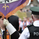 A man is wearing an orange sash and a black bowler hat and holding a banner. A policeman has his back to the camera. He has a black vest on stating police in white lettering. He has a cap and a white shirt on. A parade is out of focus in the background.