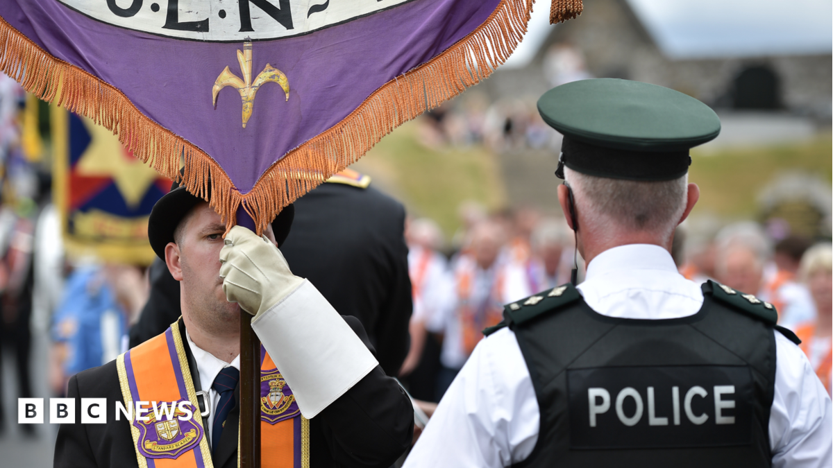 A man is wearing an orange sash and a black bowler hat and holding a banner. A policeman has his back to the camera. He has a black vest on stating police in white lettering. He has a cap and a white shirt on. A parade is out of focus in the background.