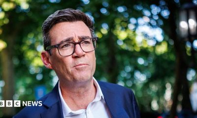 A medium close up of Andy Burnham wearing a blue suit and an open-collar white shirt with trees behind him in soft focus