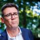 A medium close up of Andy Burnham wearing a blue suit and an open-collar white shirt with trees behind him in soft focus