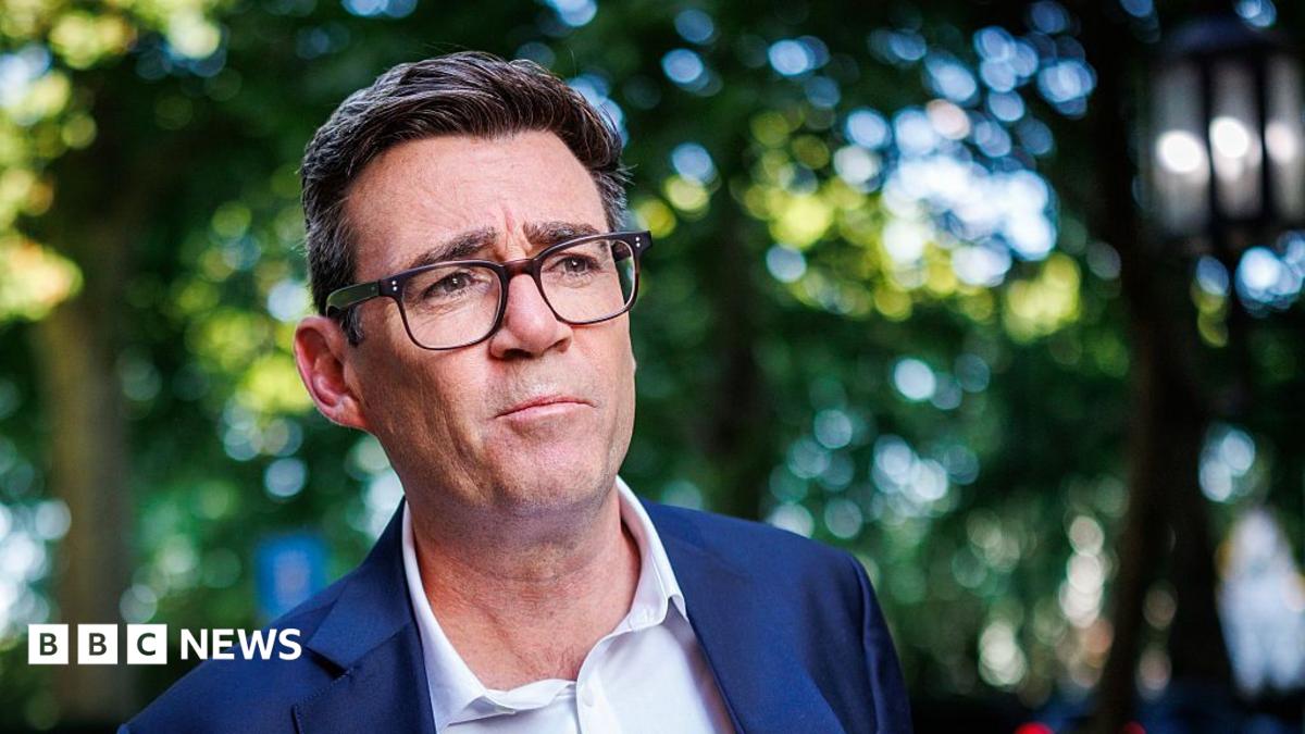 A medium close up of Andy Burnham wearing a blue suit and an open-collar white shirt with trees behind him in soft focus