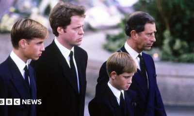 A young Prince William stands next to Earl Spencer, a young Prince Harry and Charles at Diana's funeral. They are all wearing dark suits with ties and are facing ahead solemnly.