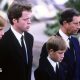 A young Prince William stands next to Earl Spencer, a young Prince Harry and Charles at Diana's funeral. They are all wearing dark suits with ties and are facing ahead solemnly.