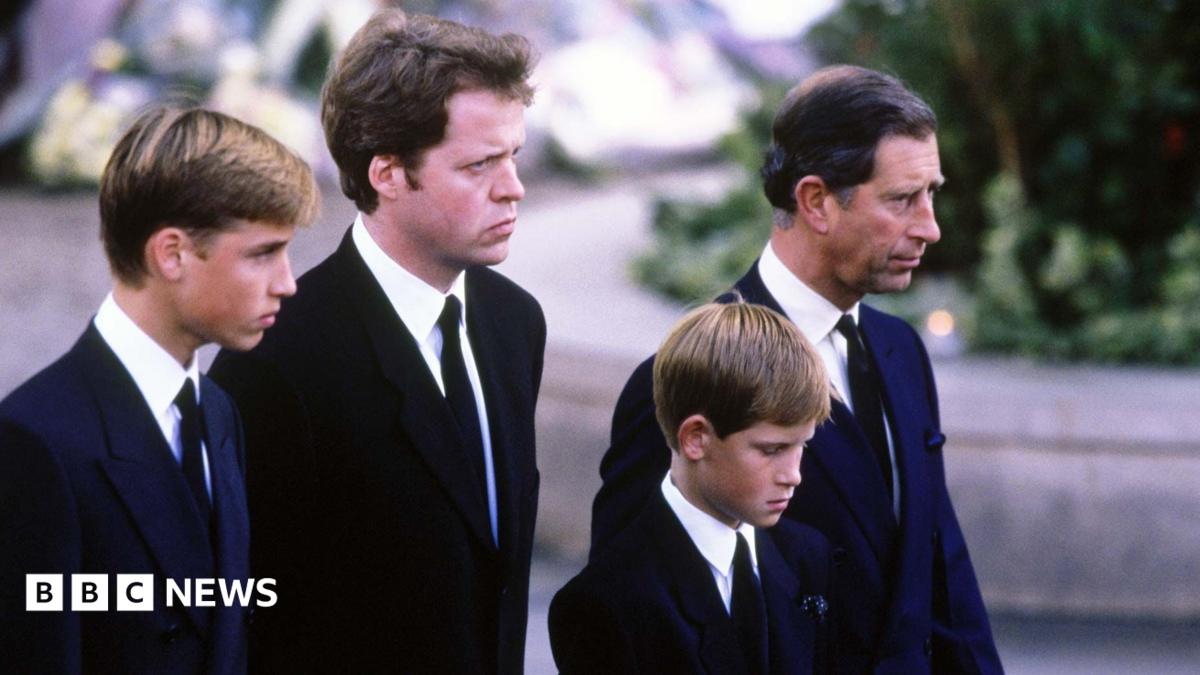 A young Prince William stands next to Earl Spencer, a young Prince Harry and Charles at Diana's funeral. They are all wearing dark suits with ties and are facing ahead solemnly.