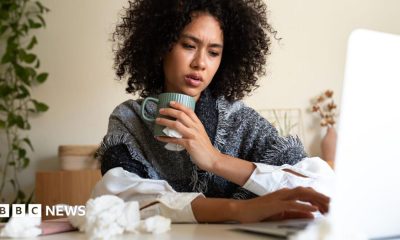 A woman wrapped in a blanket and holding a hot drink sits at a table with a laptop on it. The table has scrunched-up tissues on it as well.