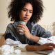A woman wrapped in a blanket and holding a hot drink sits at a table with a laptop on it. The table has scrunched-up tissues on it as well.