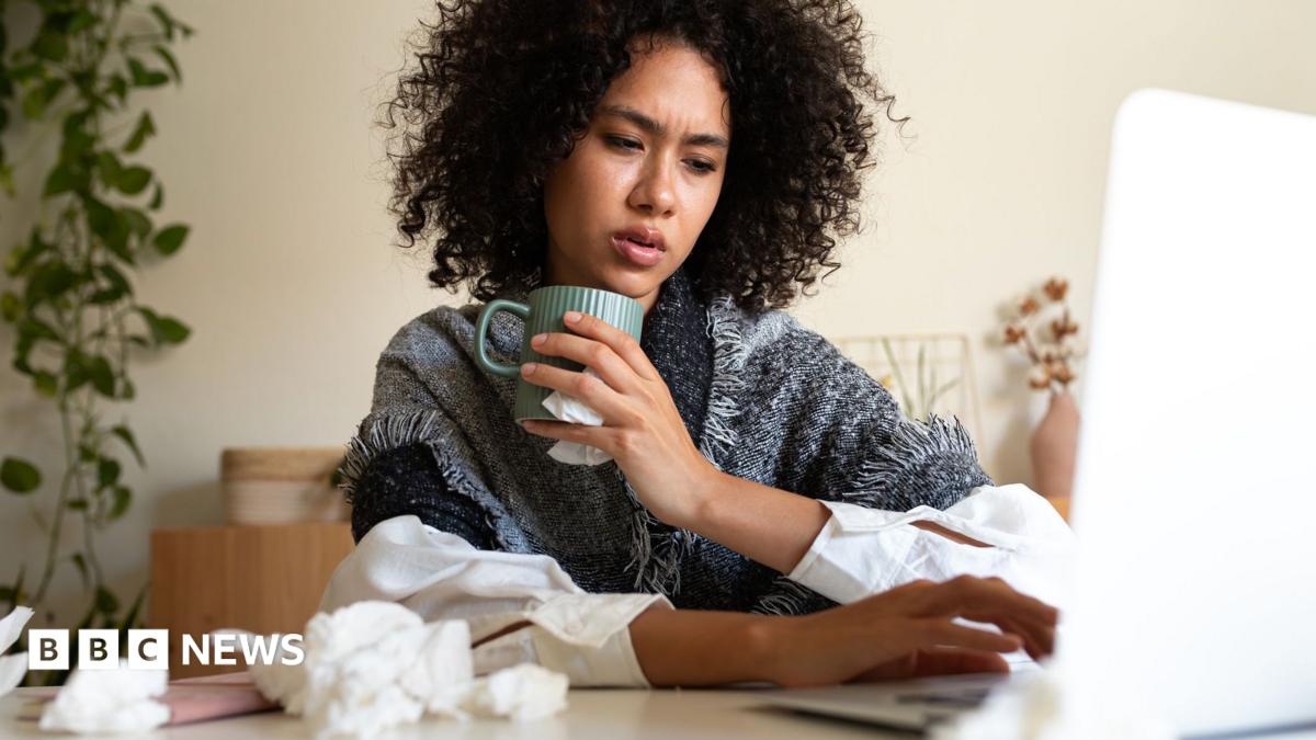 A woman wrapped in a blanket and holding a hot drink sits at a table with a laptop on it. The table has scrunched-up tissues on it as well.