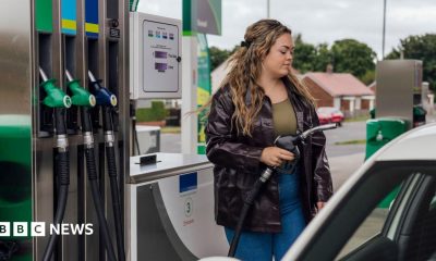 A stock image showing a woman wearing blue jeans, a green top and a black leather jacket. She has long and wavy brown hair and is filling up her car at a petrol station.