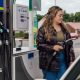 A stock image showing a woman wearing blue jeans, a green top and a black leather jacket. She has long and wavy brown hair and is filling up her car at a petrol station.