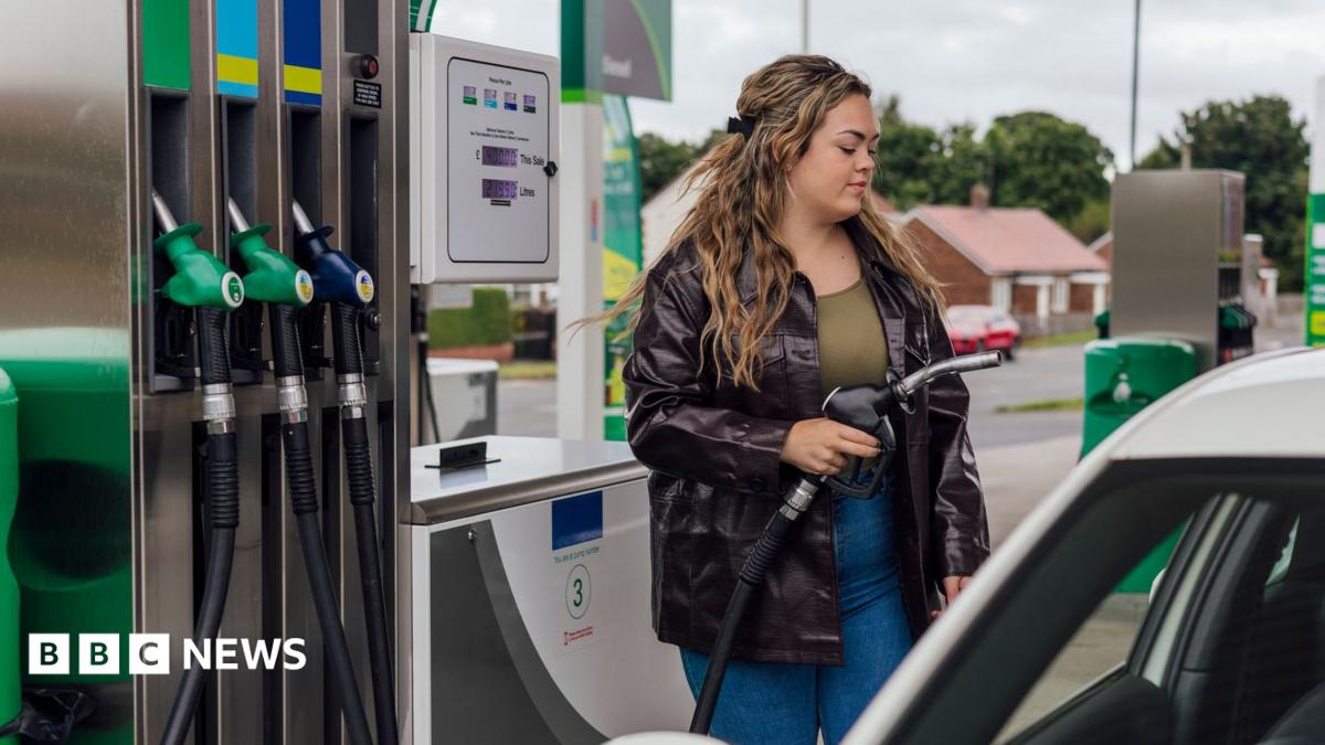 A stock image showing a woman wearing blue jeans, a green top and a black leather jacket. She has long and wavy brown hair and is filling up her car at a petrol station.