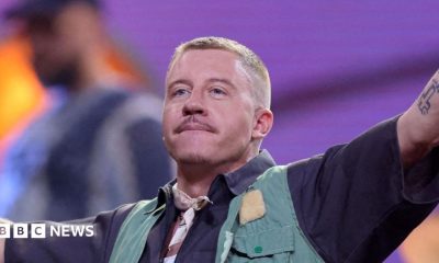 Close up of Macklemore, who has shaved blonde hair, and is wearing a blue striped shirt.